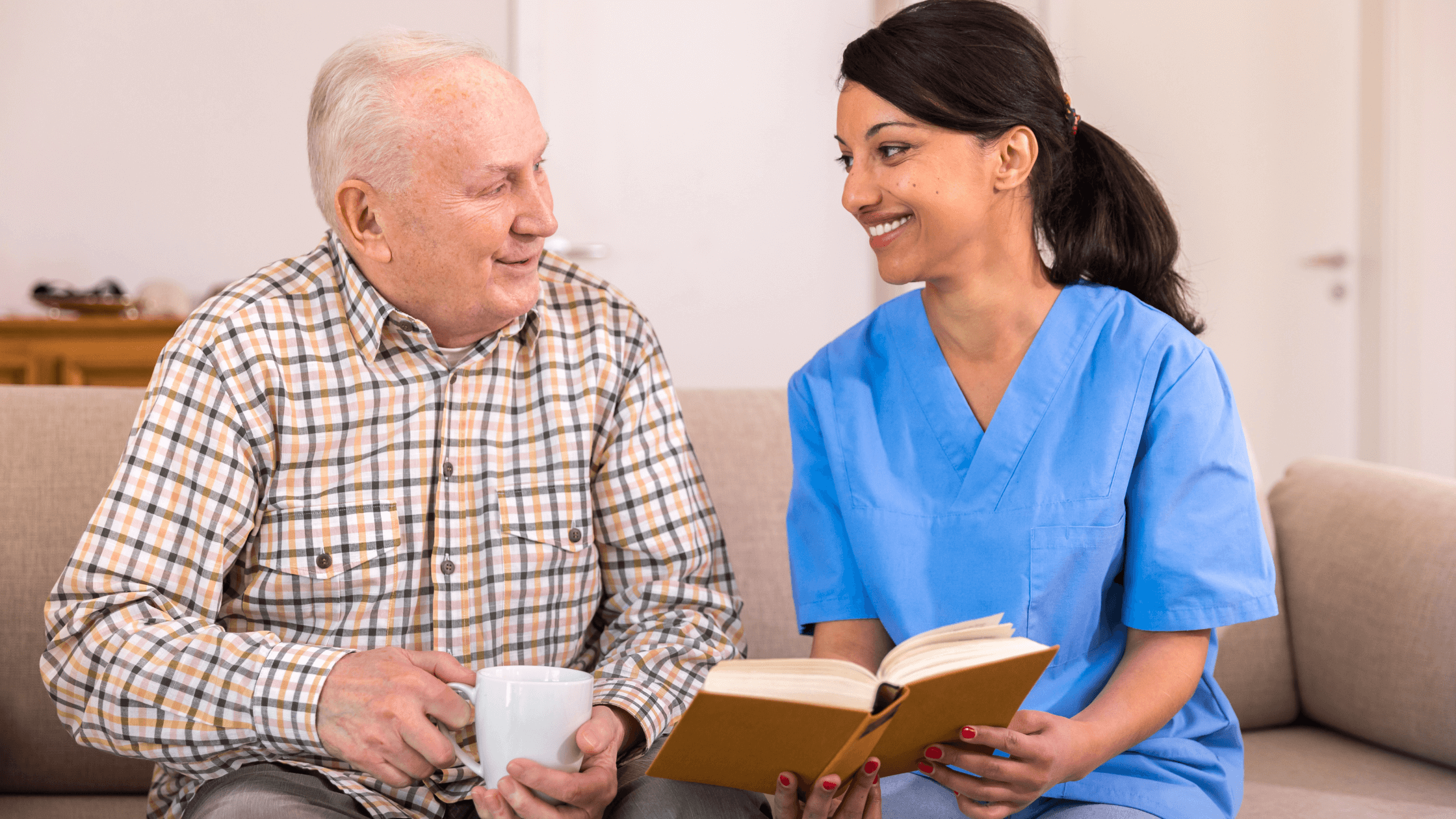 Female caregiver on uniform holding a book with senior male patient holding mug sitting on couch