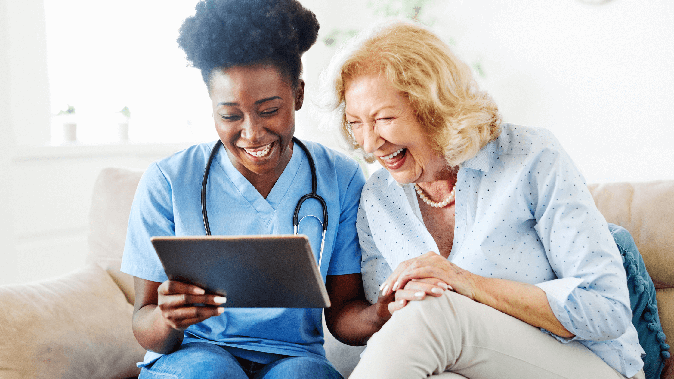 Woman of color caregiver holding a tablet with senior woman sitting beside on couch