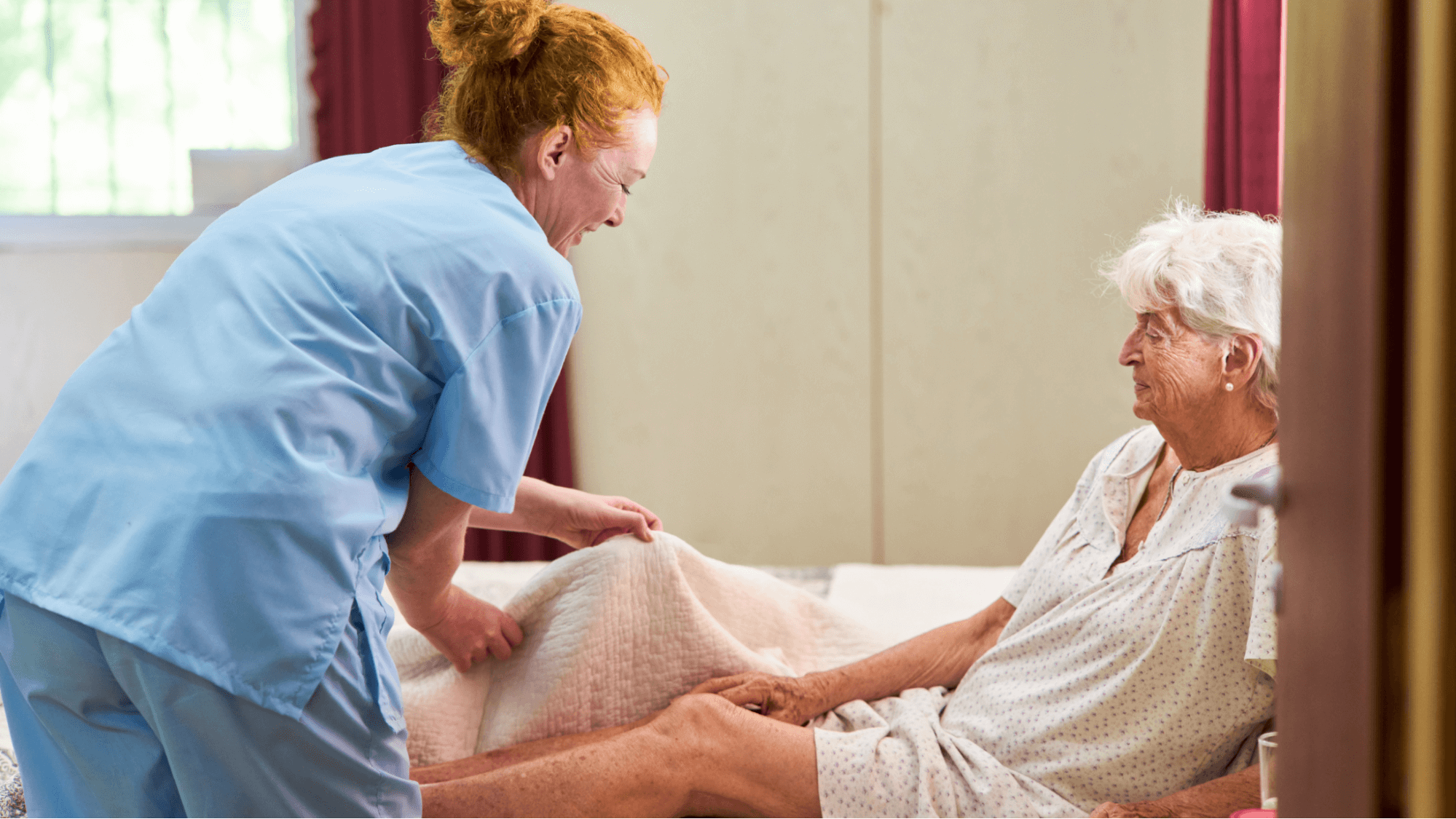 Female caregiver wearing uniform assisting senior woman on bed holding bed sheet